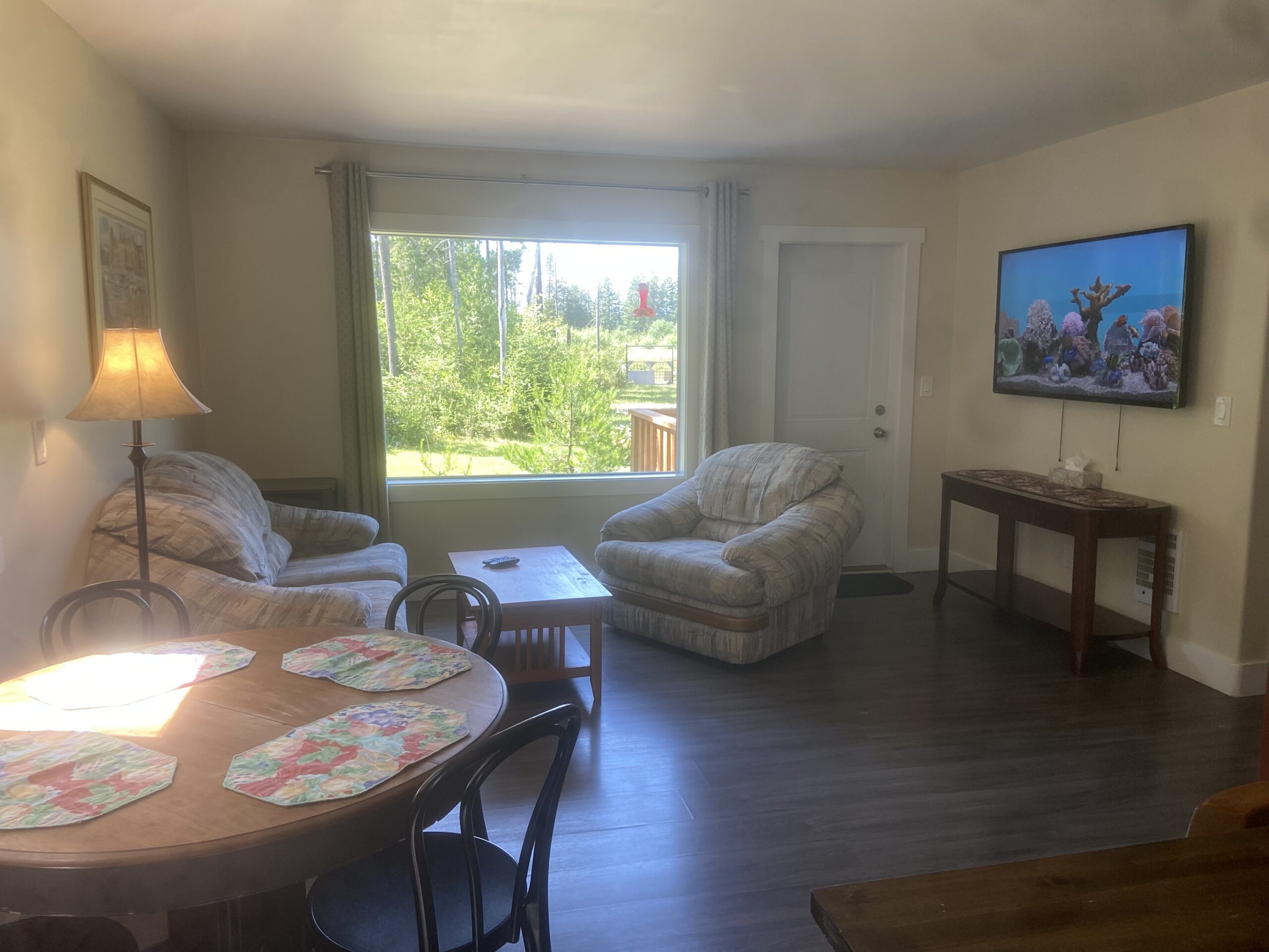 A view of the living room, a very large window, TV, couch, and loveseat are present. The dining room table is visible in the foreground